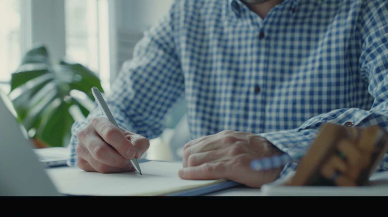 Man in plaid shirt writing notes at white desk